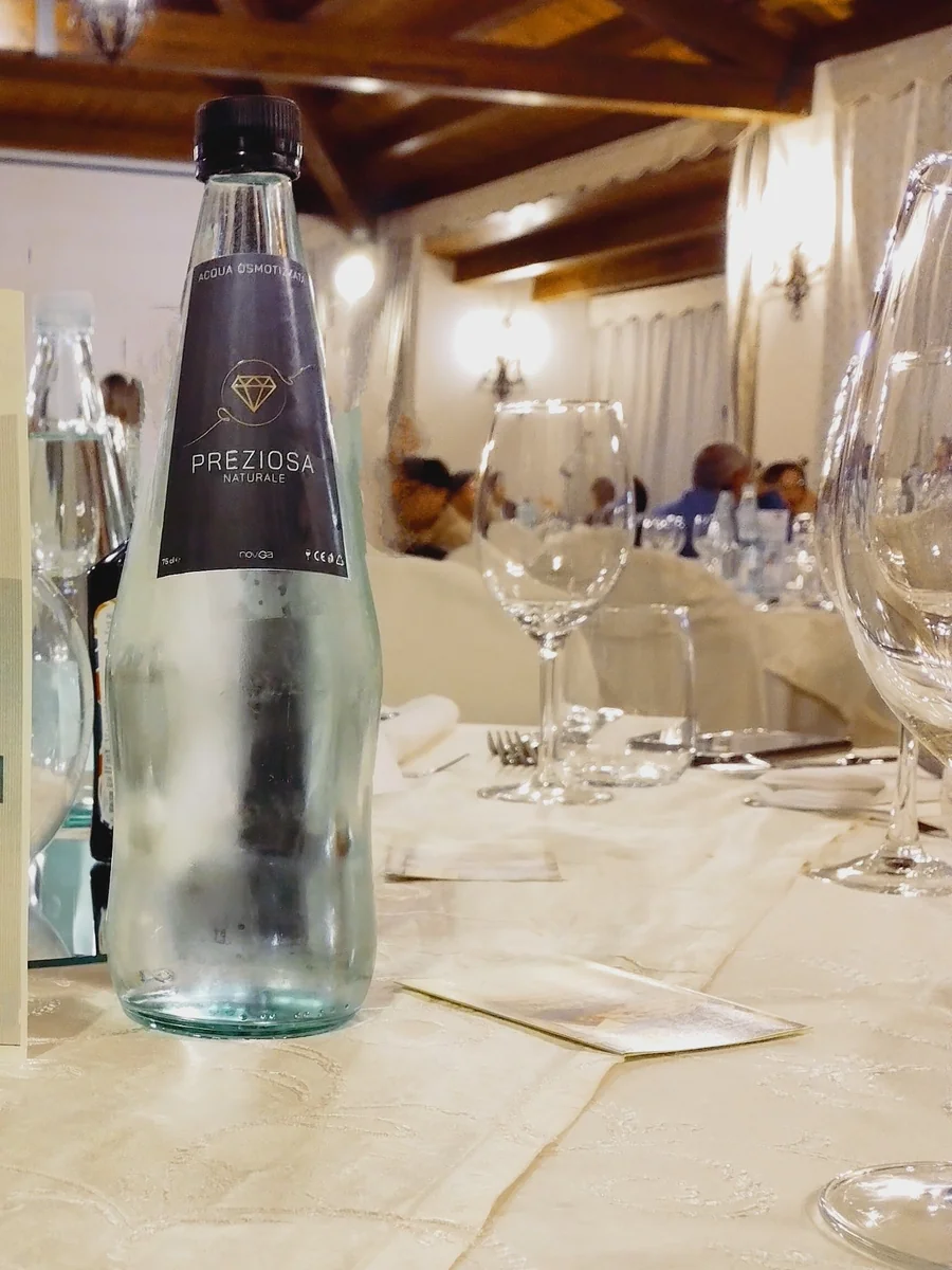 Bottiglia Preziosa Noviga con acqua osmotizzata servita in un ristorante siciliano, con bicchieri e tavolata sullo sfondo